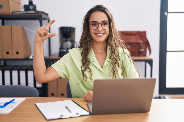 Jeune femme hispanique travaillant au bureau portant des lunettes geste souriant et confiant avec la main faisant signe de petite taille avec les doigts regardant et la caméra. concept de mesure.  - Photo, image