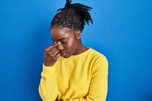 Beautiful black woman standing over blue background tired rubbing nose and eyes feeling fatigue and headache. stress and frustration concept.  - Fotó, kép