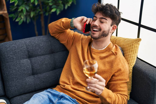 Young hispanic man talking on the smartphone and drinking wine at home - Zdjęcie, obraz