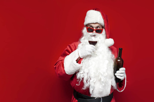 dronken santa claus in hoed en feestelijke glazen drinkt wijn uit glas op rode achtergrond, mannelijke sommelier in kerstman kostuum smaakt en drinkt alcohol voor kerst - Foto, afbeelding