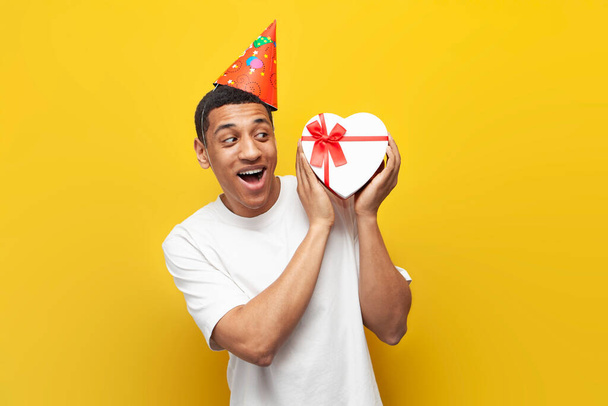 joyful african american guy in party hat celebrates birthday and holds gift box in the shape of heart, interested man with gift for valentine's day on yellow isolated background - Zdjęcie, obraz
