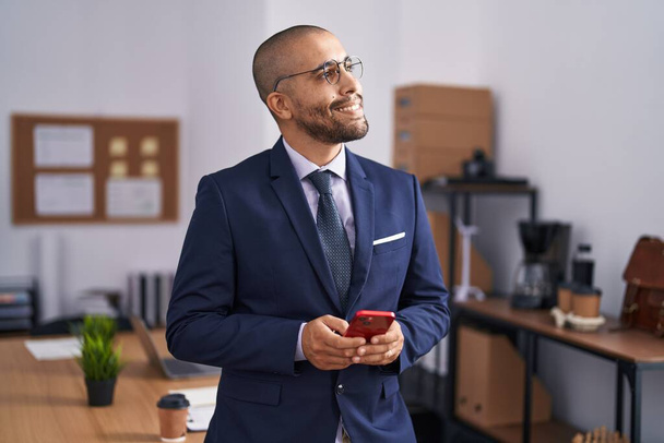 Trabajador de negocios joven latino sonriendo confiado usando teléfono inteligente en la oficina - Foto, Imagen