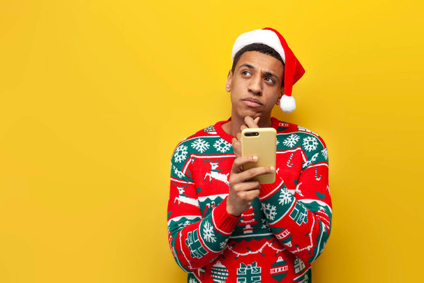 african american pensive guy in new year clothes and santa hat uses smartphone and thinks on yellow isolated background, man in sweater types online on the phone and dreams for the new year - Photo, Image