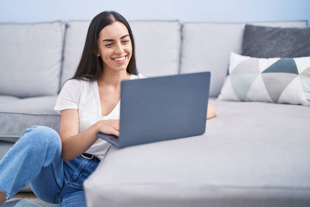 Молода іспаномовна жінка використовує ноутбук, сидячи на підлозі вдома
 - Фото, зображення