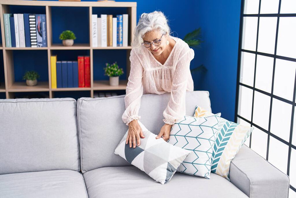 Middelbare leeftijd grijs-harige vrouw glimlachend zelfverzekerd organiseren sofa thuis - Foto, afbeelding