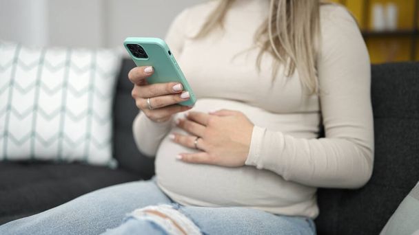 Молодая беременная женщина с помощью смартфона трогает живот дома - Фото, изображение