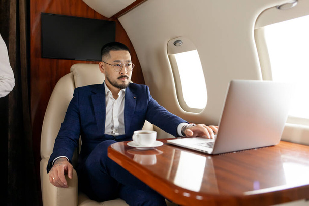 exitoso empresario asiático en traje y gafas vuela en avión privado de lujo con portátil, comerciante coreano financiero trabaja en el ordenador en avión, estilo de vida de lujo - Foto, Imagen