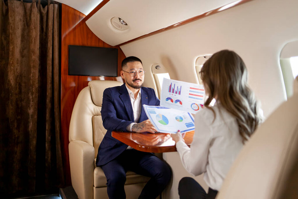 スーツと眼鏡の成功したアジアのビジネスマンは秘書補佐官と民間の高級ジェットで飛ぶ、女性マネージャーは飛行機のボスに文書や報告書を示しています - 写真・画像