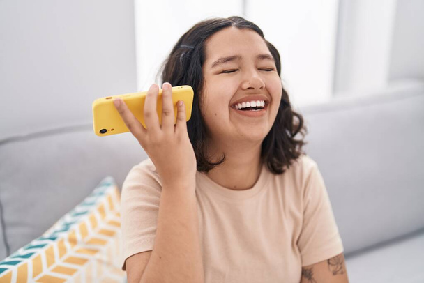 Молодая женщина прослушивает голосовое сообщение на смартфоне, сидя дома на диване - Фото, изображение