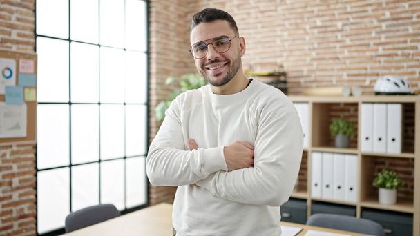 Mladý hispánec muž obchodní pracovník s úsměvem sebevědomí stojící s rukama zkřížené gesto v kanceláři - Fotografie, Obrázek