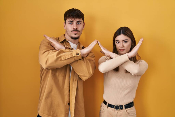 Mladý hispánský pár stojící nad žlutým odmítavým výrazem na pozadí se zkříženýma rukama dělá negativní znamení, rozzlobený obličej  - Fotografie, Obrázek