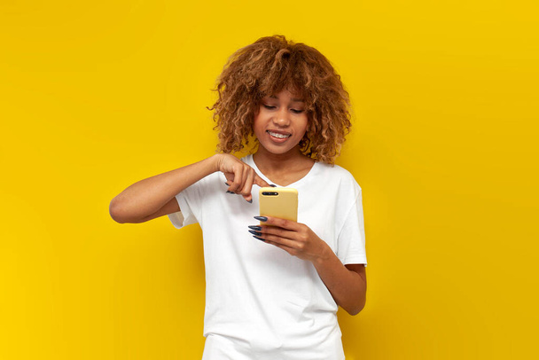 cute curly american girl with braces in white t-shirt uses smartphone and smiles on yellow isolated background, young woman is typing message and looking at the phone - Photo, Image