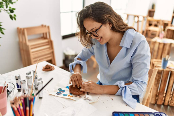 Junge schöne hispanische Künstlerin lächelt zuversichtlich machen Keramiktopf mit Händen im Kunstatelier - Foto, Bild