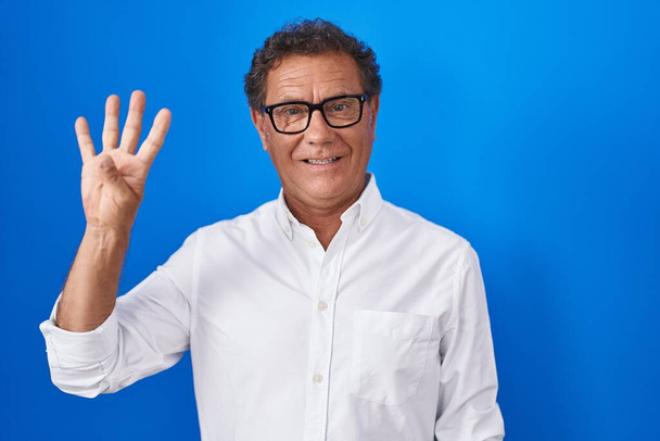 Középkorú spanyol férfi áll a kék háttér mutatja, és rámutatva ujjak száma négy, miközben mosolygós magabiztos és boldog.  - Fotó, kép