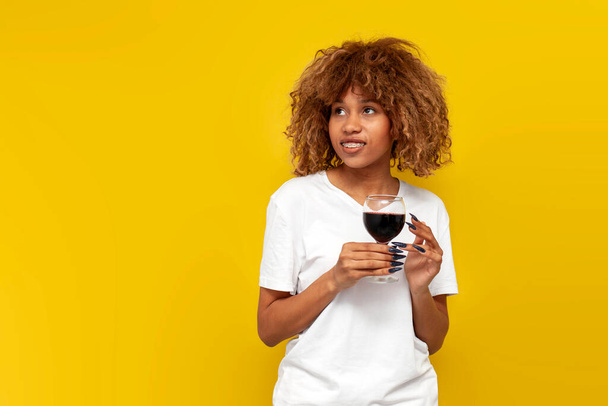 young curly american girl with braces holds glass of wine on yellow isolated background, african woman in white t-shirt with red wine smiles and looks at copy space - Photo, Image