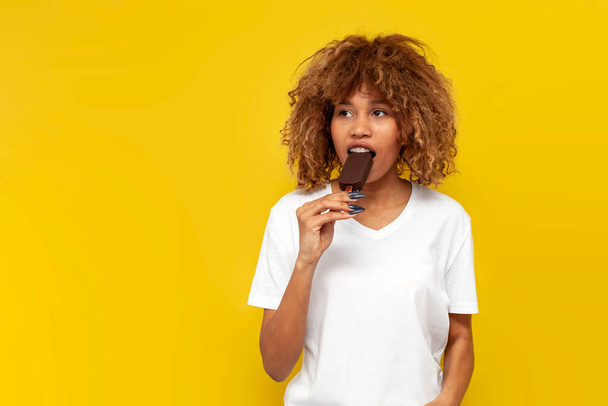 giovane ragazza riccia americana con bretelle mangia gelato al cioccolato su sfondo giallo isolato, donna africana in t-shirt bianca morde dolce dessert freddo - Foto, immagini