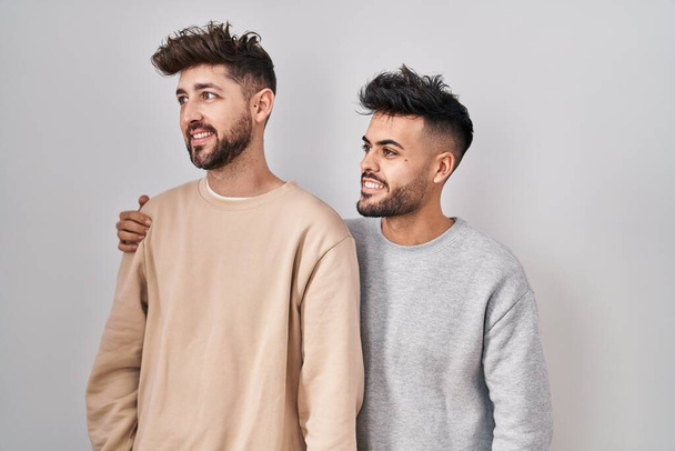 Mladý homosexuální pár stojící nad bílým pozadím, dívající se stranou s úsměvem na tváři, přirozený výraz. jistota smíchu.  - Fotografie, Obrázek