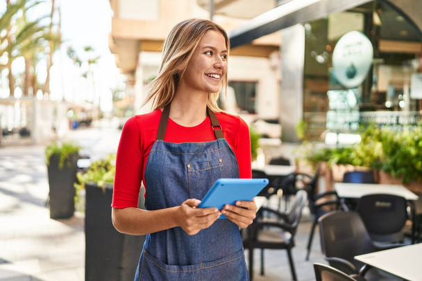Jonge Spaanse vrouw serveerster glimlachend vol vertrouwen met behulp van touchpad op coffeeshop terras - Foto, afbeelding