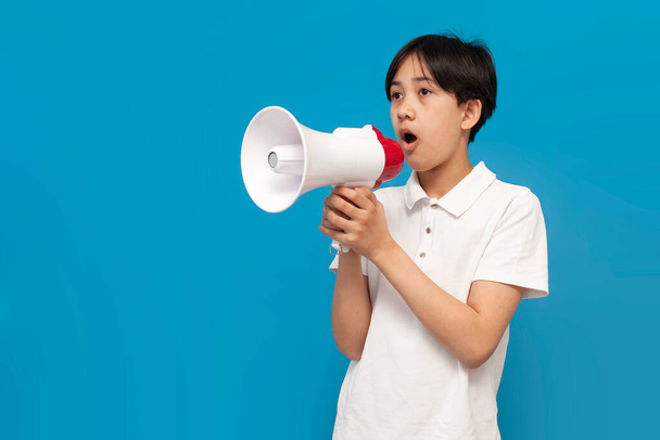 Азиатский мальчик двенадцати лет в белой футболке объявляет информацию в мегафон на синем изолированном фоне, корейский ребенок кричит в громкоговоритель - Фото, изображение