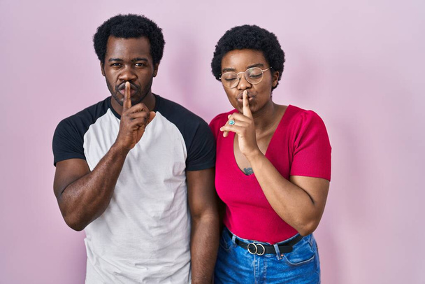 Ein junges afrikanisch-amerikanisches Paar steht vor rosafarbenem Hintergrund und bittet um Ruhe mit dem Finger auf den Lippen. Schweigen und geheimes Konzept.  - Foto, Bild