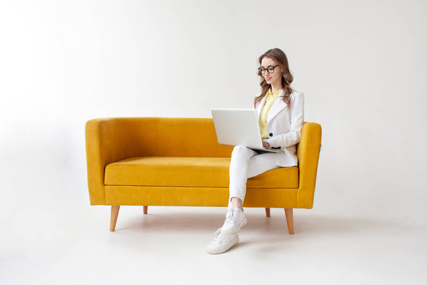 young businesswoman in suit sits on comfortable soft sofa and uses laptop, girl in formal wear is typing on computer on yellow couch on white isolated background - Foto, imagen