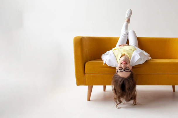 νεαρή επιχειρηματίας με κοστούμι βρίσκεται και στηρίζεται σε άνετο μαλακό καναπέ, κορίτσι με επίσημη ένδυση είναι lounging και τεμπέλης σε κίτρινο καναπέ σε λευκό απομονωμένο φόντο - Φωτογραφία, εικόνα