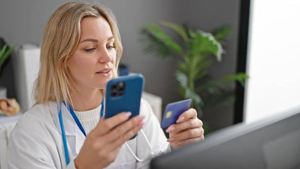 Young blonde woman doctor shopping with smartphone and credit card at clinic - Photo, Image