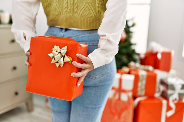 Young beautiful hispanic woman holding christmas gift on back at home - Foto, immagini