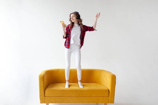 nuori söpö tyttö tanssii musiikkia ja hyppää pehmeä mukava sohva, nainen kuulokkeet kuuntelee musiikkia ja käyttää älypuhelinta keltainen sohva valkoinen eristetty tausta - Valokuva, kuva