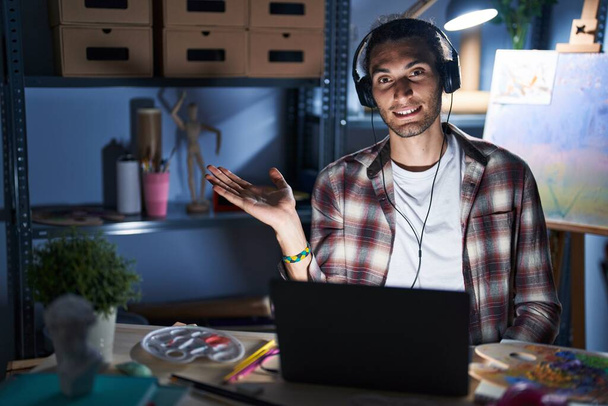 Fiatal spanyol férfi ül a művészeti stúdióban laptop késő este mosolygós vidám bemutató és mutató tenyérrel néz a kamerába.  - Fotó, kép