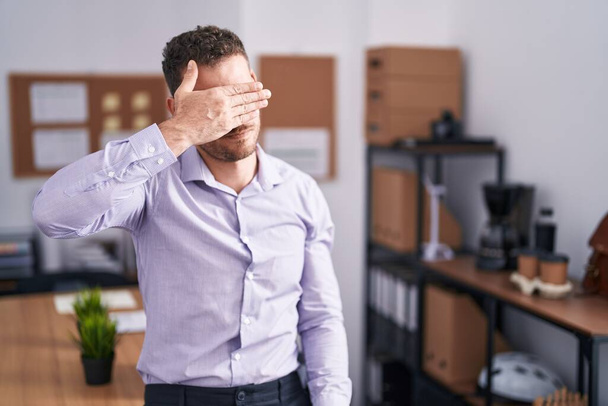 Giovane uomo ispanico in ufficio che copre gli occhi con la mano, guardando serio e triste. concetto di avvistamento, nascondiglio e rifiuto  - Foto, immagini