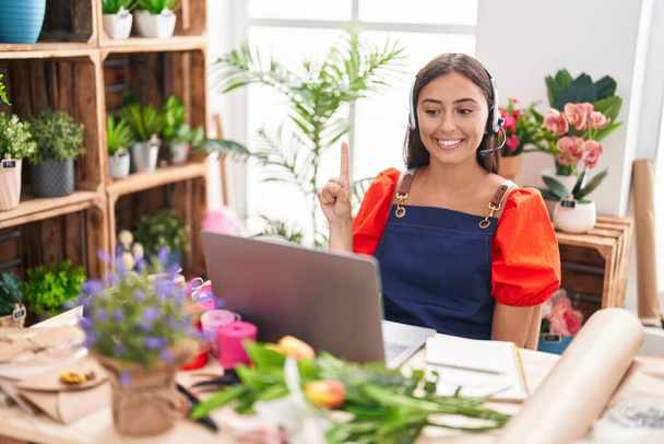 Mujer hispana joven trabajando en floristería haciendo videollamadas sonriendo con una idea o pregunta señalando con el dedo con la cara feliz, número uno  - Foto, imagen