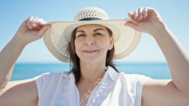 Střední věk hispánská žena turista úsměv držení letní klobouk na pláži - Fotografie, Obrázek