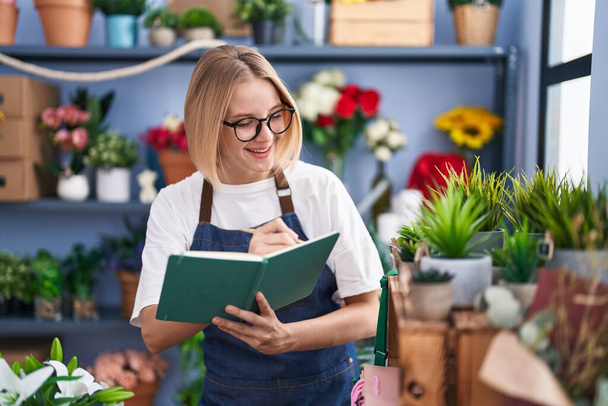 Giovane donna bionda fiorista sorridente scrittura fiduciosa sul taccuino al negozio fiorista - Foto, immagini