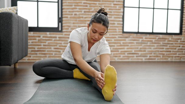 Young beautiful hispanic woman sitting on floor stretching legs at home - Photo, Image