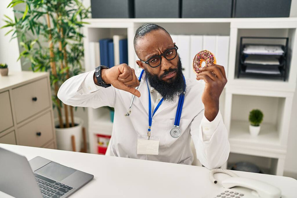 Homme afro-américain travaillant à la clinique diététiste tenant un beignet avec un visage en colère, signe négatif montrant un aversion avec les pouces baissés, concept de rejet  - Photo, image
