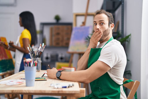 Ισπανός στο στούντιο ζωγραφικής που καλύπτει το στόμα με το χέρι, σοκαρισμένος και φοβισμένος για λάθος. έκπληκτη έκφραση  - Φωτογραφία, εικόνα