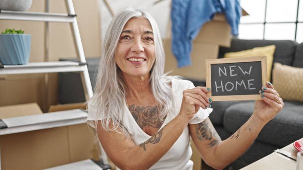 Moyen âge femme aux cheveux gris souriant confiant tenant tableau noir à la nouvelle maison - Photo, image