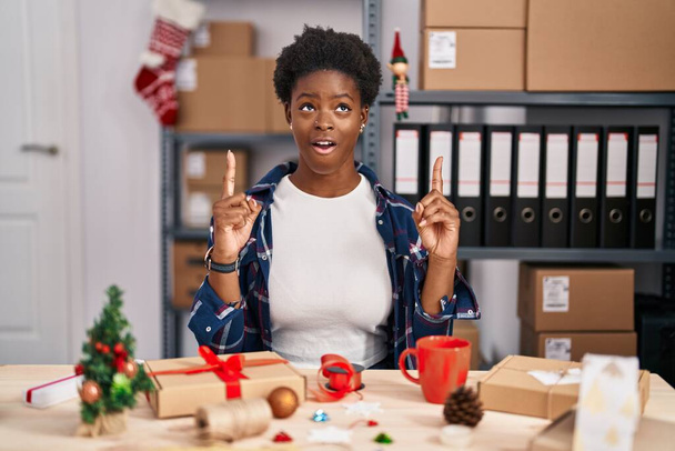 Afrikkalainen amerikkalainen nainen työskentelee pienissä yrityksissä tehden joulukoristeita hämmästynyt ja yllättynyt katselee ylös ja osoittaa sormilla ja nosti kädet.  - Valokuva, kuva