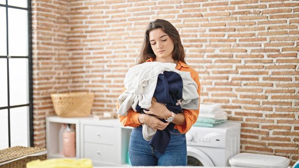 Young beautiful hispanic woman holding stack of clothes at laundry room - 写真・画像