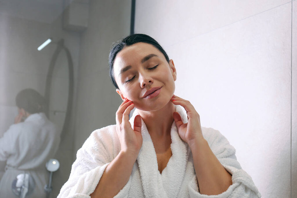 Young woman wearing a bathrobe looking at the round shaped mirror and applying anti-aging skincare to her face with massaging motions. Morning beauty routine concept. Close up, copy space, background. - Φωτογραφία, εικόνα