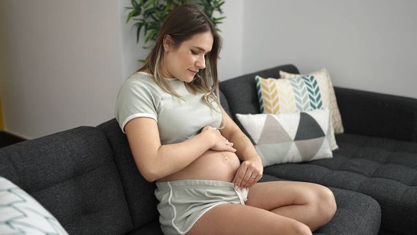 Mujer embarazada joven tocando el vientre con expresión relajada en casa - Foto, Imagen