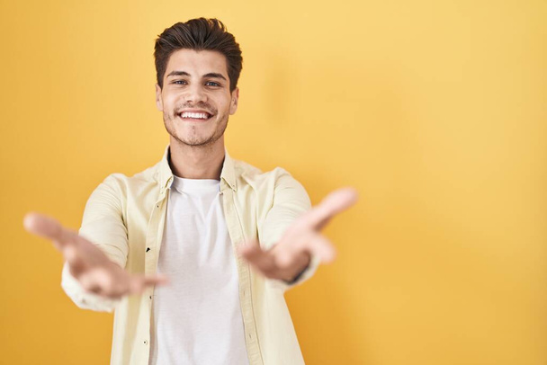 Молодий іспаномовний чоловік стоїть над жовтим тлом усміхнений веселий, що пропонує руки, що допомагають і приймають
.  - Фото, зображення