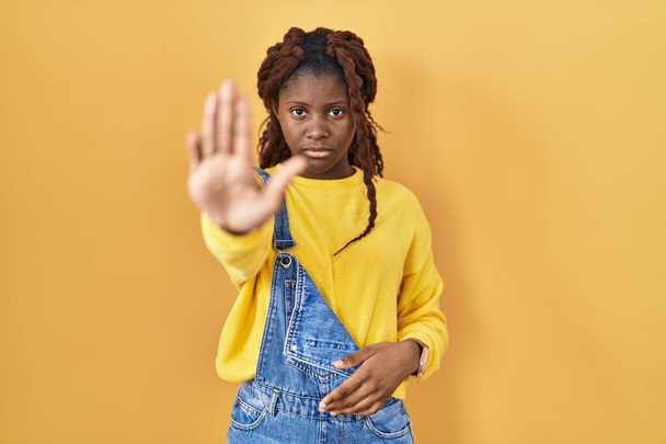 Afrikanerin, die vor gelbem Hintergrund steht und nicht mehr mit der Handfläche singt. Warnausdruck mit negativer und ernster Geste im Gesicht.  - Foto, Bild