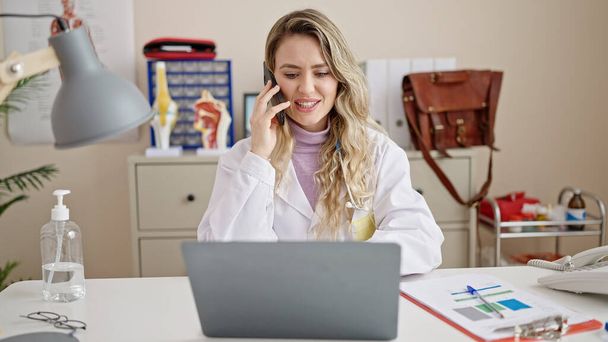Junge blonde Ärztin telefoniert in Klinik mit Laptop auf Smartphone - Foto, Bild