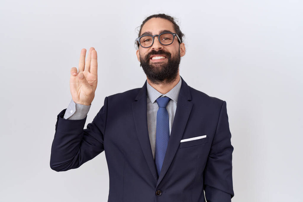 Homem hispânico com barba vestindo terno e gravata mostrando e apontando para cima com os dedos número três, sorrindo confiante e feliz.  - Foto, Imagem