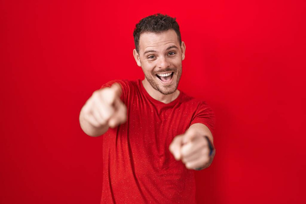 Fiatal spanyol férfi áll a piros háttér mutat rád és a kamera ujjaival, mosolygós pozitív és vidám  - Fotó, kép