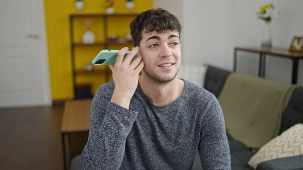 Young hispanic man listening to voice message smiling at home - 写真・画像