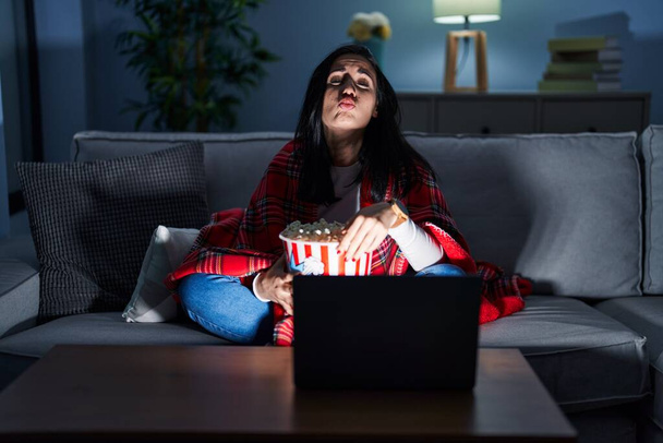 Latynoska kobieta jedząca popcorn oglądająca film na kanapie patrząca w kamerę, dmuchająca pocałunek na antenie, jest urocza i seksowna. wyrażenie miłości.  - Zdjęcie, obraz