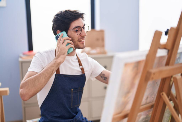 Young hispanic man artist talking on smartphone drawing at art studio - Photo, Image
